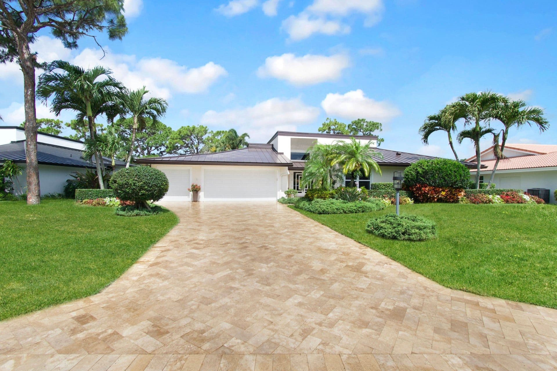 Contemporary single-story home with white exterior, flat roof, and two-car garage accessed by a paved driveway, surrounded by tropical landscaping with palm trees and lush green lawn